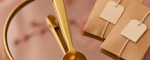 Cuillère à thé dorée posée sur un plateau miroir doré, entourée de sachets de thé kraft et de fleurs de cerisier.
